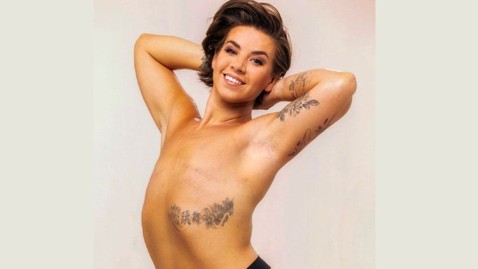 Photo of Danielle Moore for the One More Page Three campaign. She is stood against a solid, light grey background, with her arms up behind her head, showing the scars across her chest from her mastectomy. Her back is arched slightly and she is looking directly at the camera with a large smile on her face. She has two flower tattoos - one across her ribcage and another on her arm.