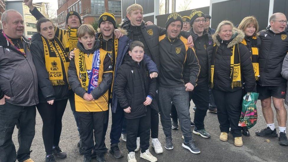 A large family group cheer and smile whilst wearing Maidstone united outfits and hats outside the stadium