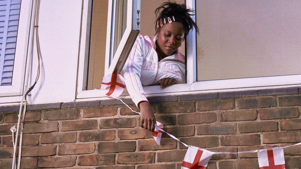Rachel Chinouriri hangs England flag bunting outside a window. Rachel is a 25-year-old black woman with long dark hair worn tied back with her side fringe secured with five multi-coloured clips. She wears a pink and white Lonsdale tracksuit top and leans out of an open window as she secured the bunting.