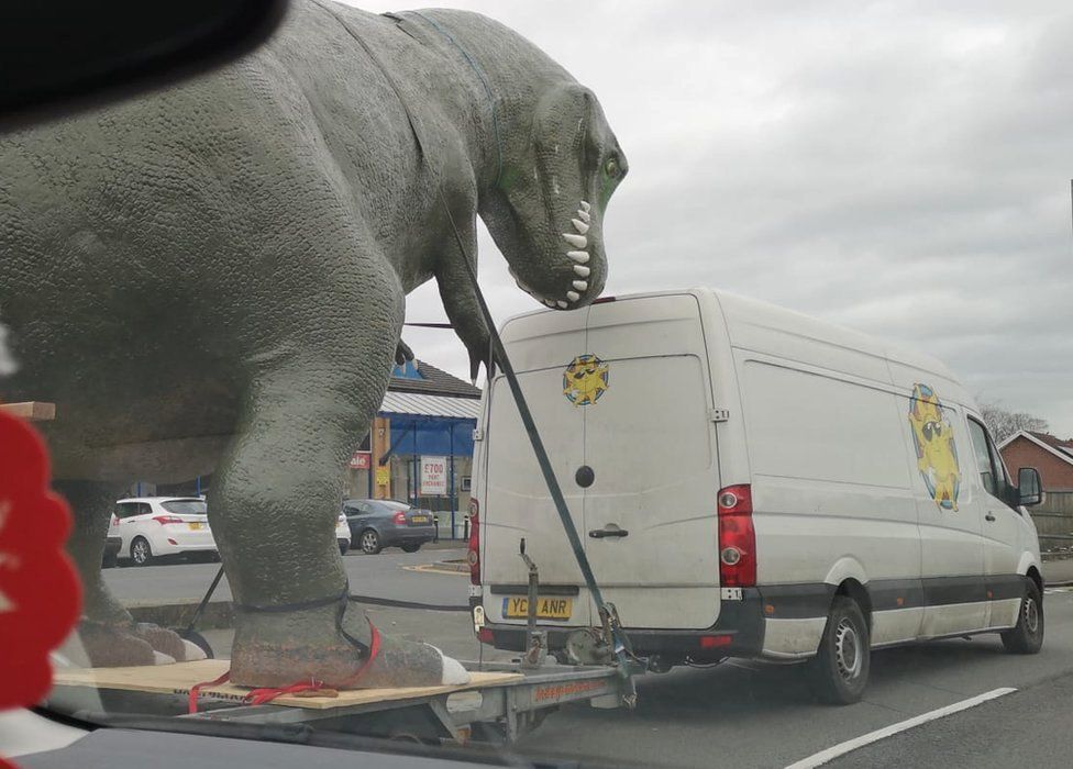 The dinosaur on the back of the trailer on the road