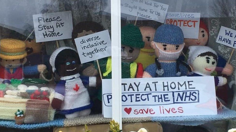 Knitted figures in window with signs "Stay at home. Protect the NHS" and "We stayed at work for you now stay at home for us please"