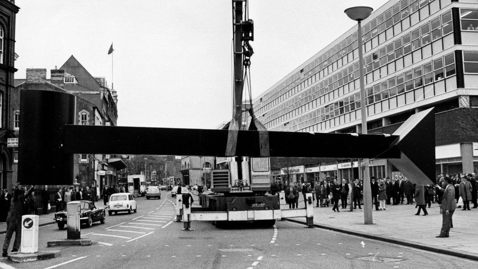 A black and white photograph of the sculpture being lifted by a crane