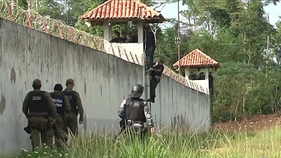 Multiple prison officers stand in a watchtower looking in to the prison, while from our ground level view we can see three more walk around the wall beneath- all heavily armoured