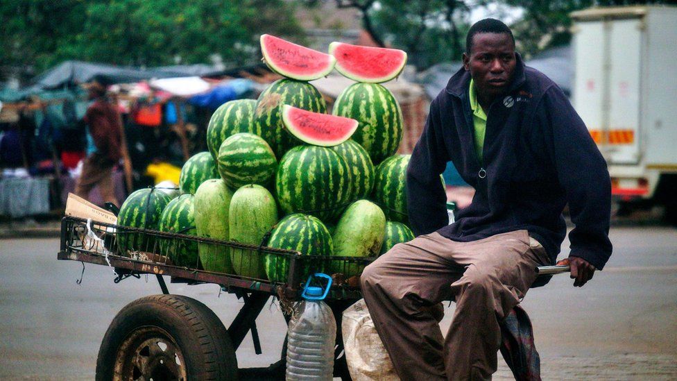 Watermelons are piled high on a cart, with few cut slices on display on top, as the seller sits on the cart's arms amid a busy marketplace