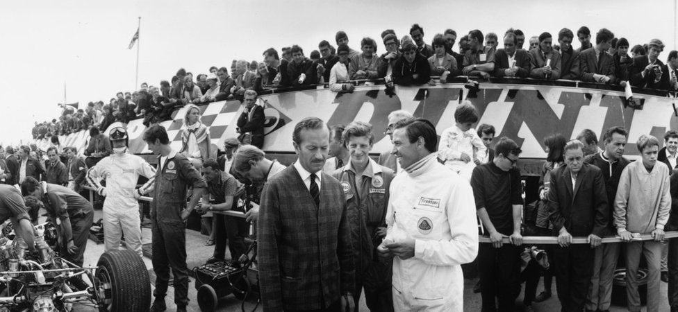 14th July 1967: During a pit stop before a practice lap at Silverstone, racing driver Jim Clark (1936 - 1968) on right in white. talks with racing boss Colin Chapman (1928 - 1981) founder of Lotus cars. Figure in racing gear on the left talking to a mechanic is racing driver Graham Hill (1929 - 1975)