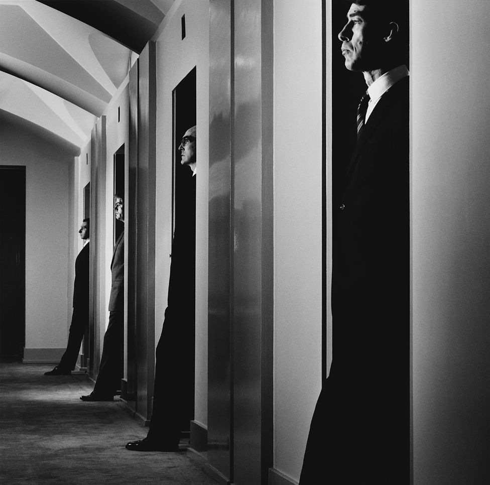 Men pose in doorways in an office corridor