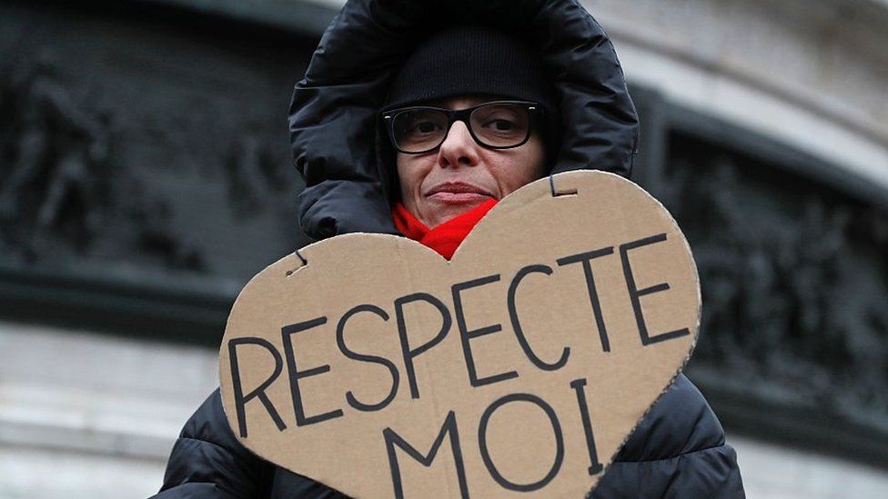 Woman in hooded jacket holds a cardboard with the message "respect me" in French