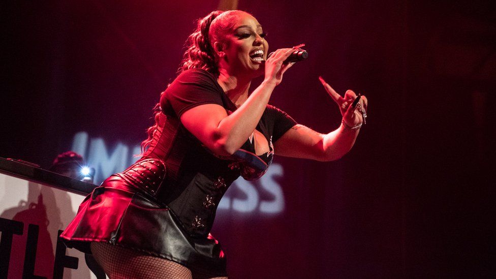 Miss LaFamilia performs at The Roundhouse on April 16, 2022 in London. The singer, a young black woman, is wearing a black leather mini skirt over fish net tights with a black corset and black Iron Maiden band T-shirt. She has her hair tied back in a long wavy pony tail and leans forward as she leans to sing into the microphone which she holds in her right hand. Her left arm is also raised with her index finger pointing up. The Roundhouse venue is dark and Miss La Familia is lit by a red spotlight.