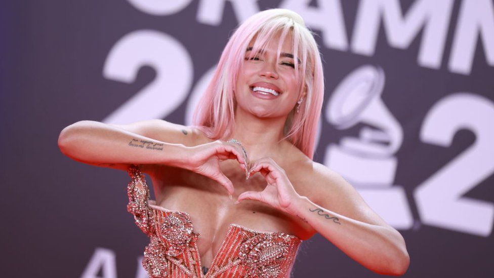 Karol G doing a heart sign with her hands, with pink hair and a pink sparkly dress