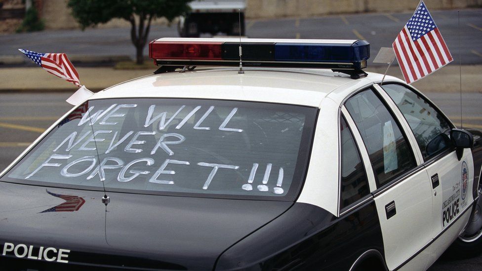 One month after the Oklahoma City bombing, a police car carries American flags and the words, "We Will Never Forget!!!"