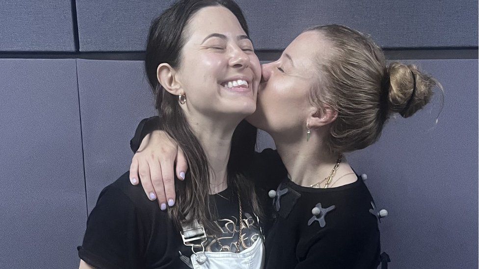 One woman in dungarees smiles while another woman wearing a motion capture suit kisses her on the cheek. They look happy and comfortable in each other's company - it's a warm, affectionate scene.