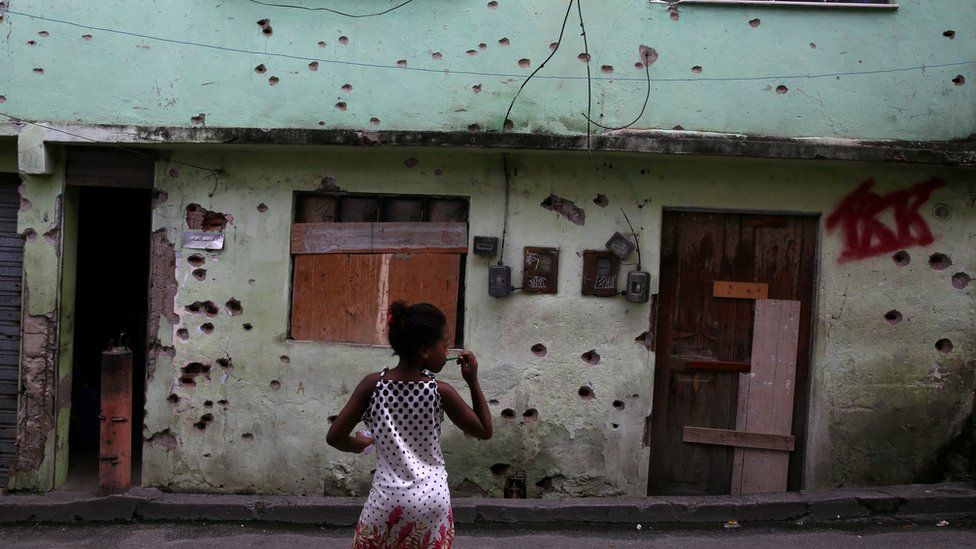 A girl stands in front of a house which is damaged by bullet holes in the Complexo de Alemao favela in Rio de Janeiro, Brazil