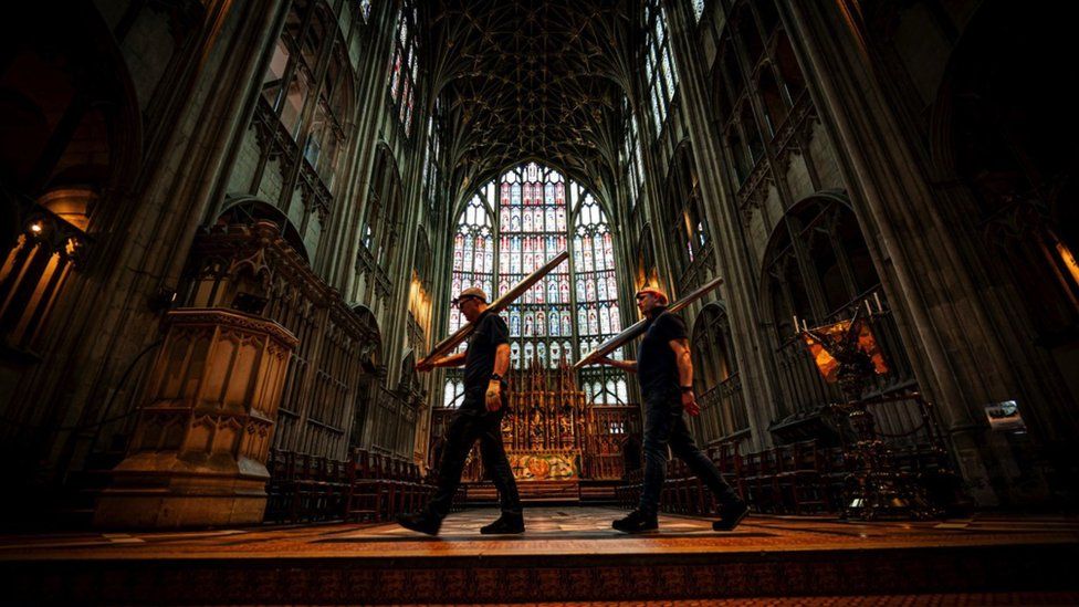 Two men walking through Gloucester Cathedral. They are both wearing hard hats and carrying equipment.