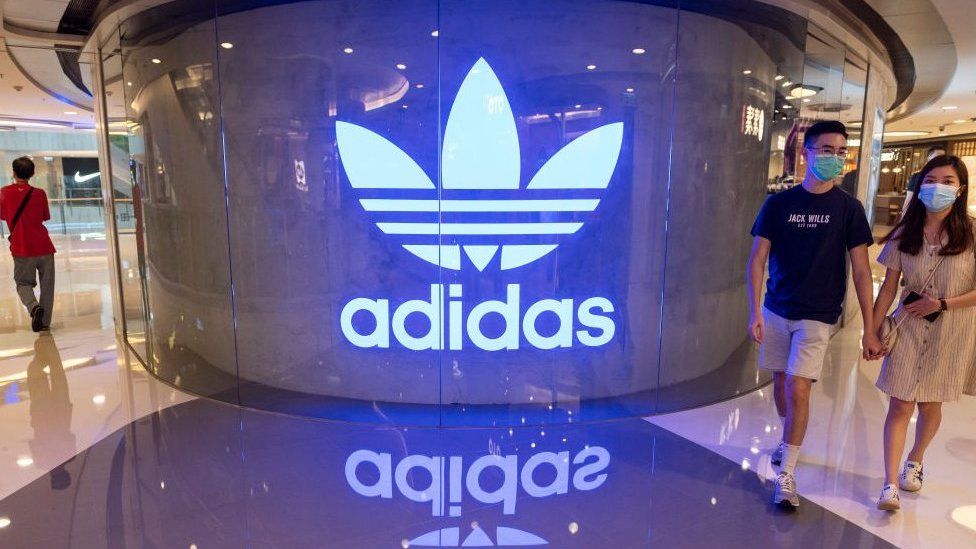 Shoppers wearing face masks walk past an Adidas logo at a shopping mall.