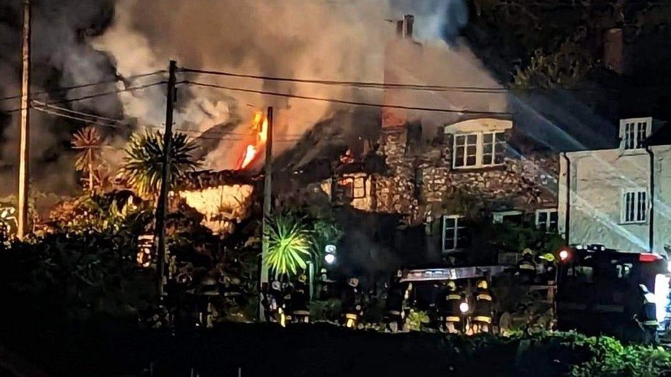 Two thatched cottages on fire at night with firefighters in the foreground