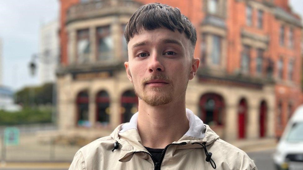 William Greenhalgh is a 21-year-old white man with short brown hair. He has a fair beard and dark blue/ grey eyes. He wears a cream coloured waterproof over a black T-shirt and looks at the camera with a neutral expression. He is photographed outside in Birmingham, behind him is a Victorian-style redbrick building.