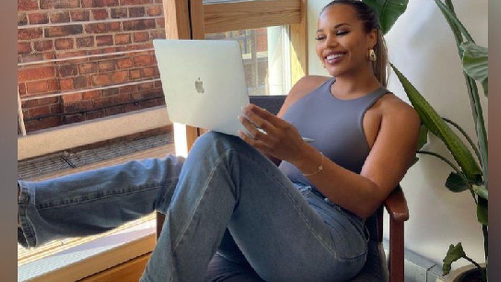Lateysha, who starred in Big Brother in 2016, sits in a chair smiling and holding a laptop