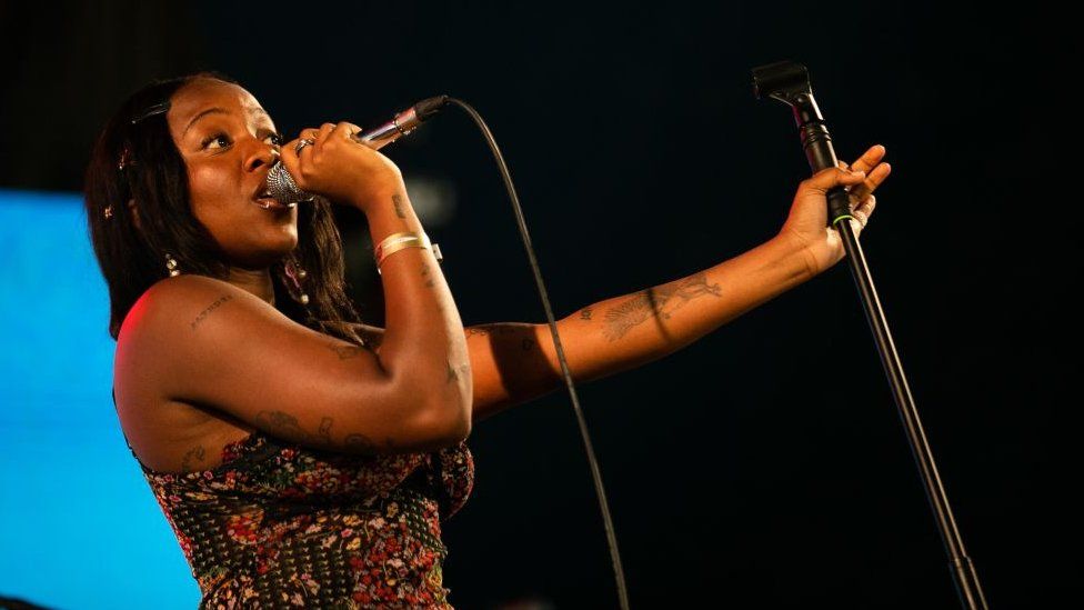 Rachel Chinouriri performing in 2023. Rachel is a black woman in her 20s with shoulder-length hair. She wears a strappy patterned top and holds a microphone to her face with her right hand, revealing small tattoos on her arms. She holds the mic stand with her left hand, outstretched from her. The staging behind her is dark, with a blue square.