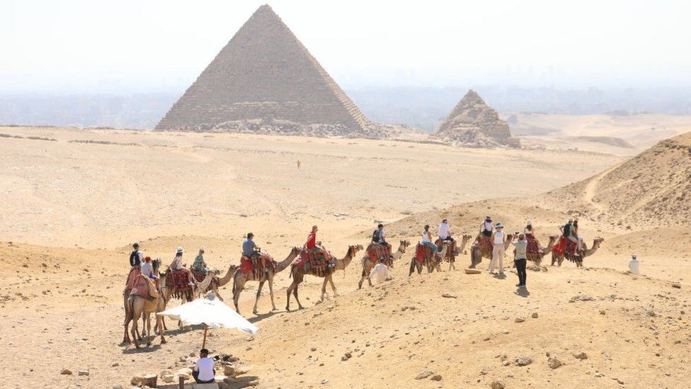 Two pyramids in the Egyptian surrounded by acres of sandy desert are visible in the background behind a haze of heat. The one on the left is very large, while the structure to its right is much smaller. In the foreground, a line of camels carrying tourists towards the attraction snakes across the frame. One couple appears to have dismounted, with a woman in a sunhat and white vest posing for a photo taken by her partner. In the bottom left corner of the frame a man shelters from the bright sun under a white parasol on a post driven into the sand.
