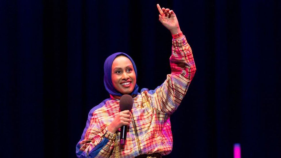 Ola Labib performing at BBC 1Xtra Comedy Gala at the Hackney Empire on 1 April 2024. Ola is a British Sudanese woman in her 30s. She has brown eyes and smiles, wearing a blue hijab and a checked-patterned tracksuit in yellow, red and blue. She holds a microphone in her right hand while pointing to the ceiling with her left. The staging behind her is black with pink accents.
