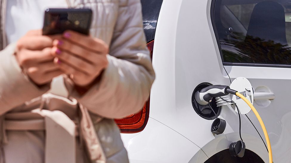 Person using smartphone with electric car charging in background