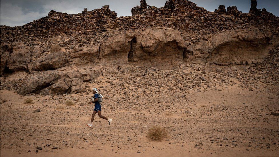 A man running in the Sahara. Most of the photo shows the arid desert in the background with a rocky landscape and it is sandy all over.