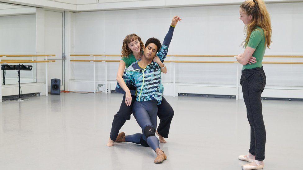 Choreographer Cathy Marston behind Marcelino Sambé, shows Lauren Cuthbertson how to hold her cello during rehearsal