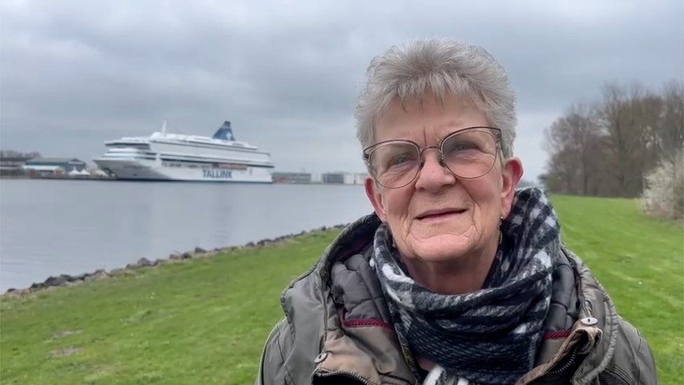Gerry, a woman with short grey hair and glasses, pictured on the river bank with the ferry in the background