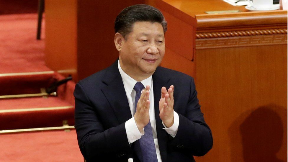 Xi Jinping, clapping, with tea and a notepad and pen on the desk in front of him