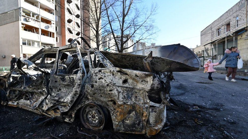 A woman and her child walk past a car destroyed by shelling in the outskirts of Ukraine's capital, Kyiv (28 February 2022)