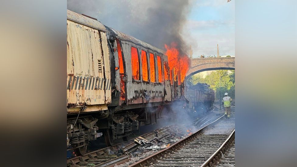 A railway track and a train carriage on fire with smoke pouring out of it