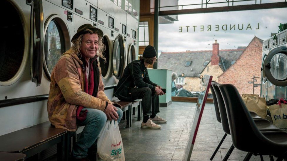 A man sits in a launderette and smiles past the camera