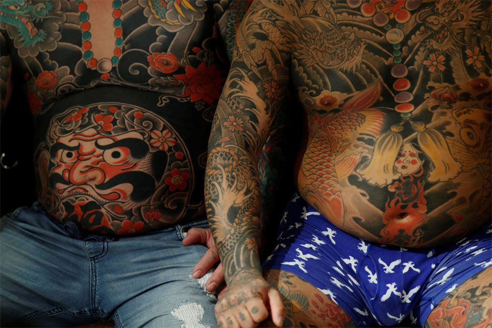 Two men sit next to each other with heavily tattooed torsos