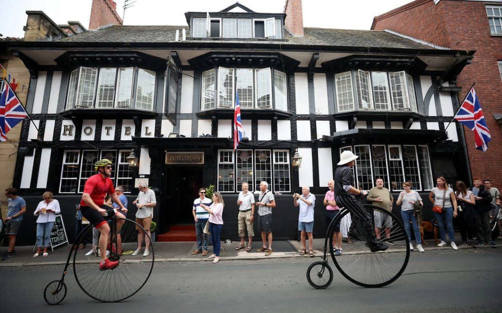 two men ride past black and white timber building and spectators