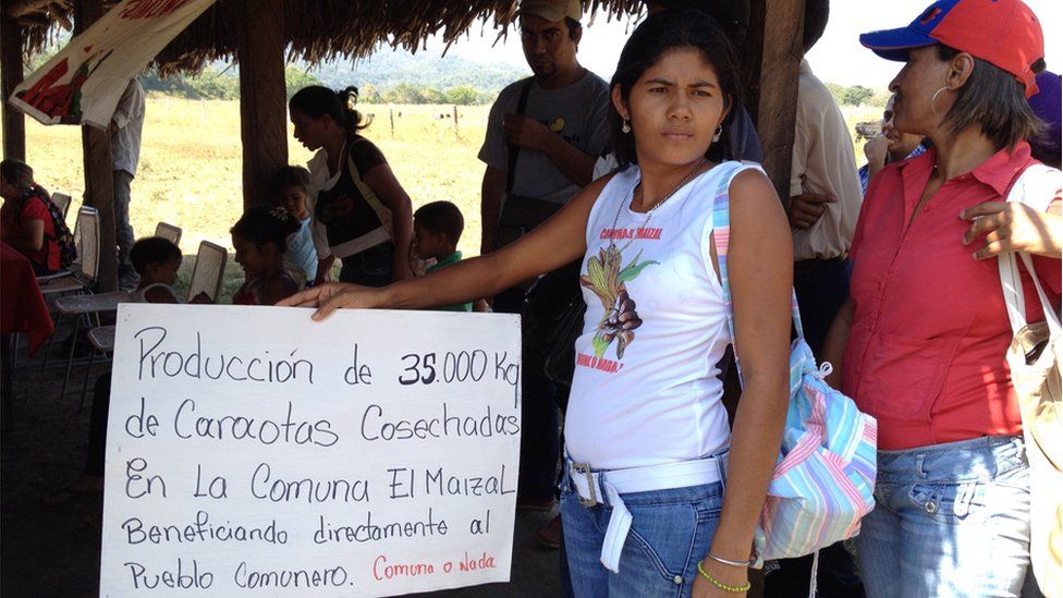 A woman at a meeting in El Maizal holds up a sign reading "Production of 35,000 kilos of sown black beans in Commune El Maizal have directly benefitted the commune"