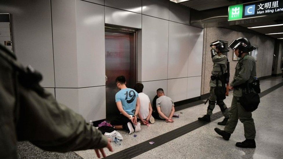 Suspected protesters kneel on the ground at a Hong Kong metro station as police guard them