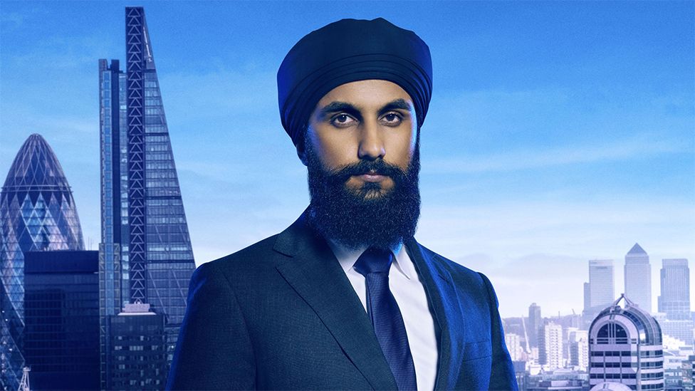 Virdi, an Asian man wearing a black turban representing his Sikh faith. He is wearing a white shirt, navy tie and dark suit, with the background of large skyscrapers in the city of London.