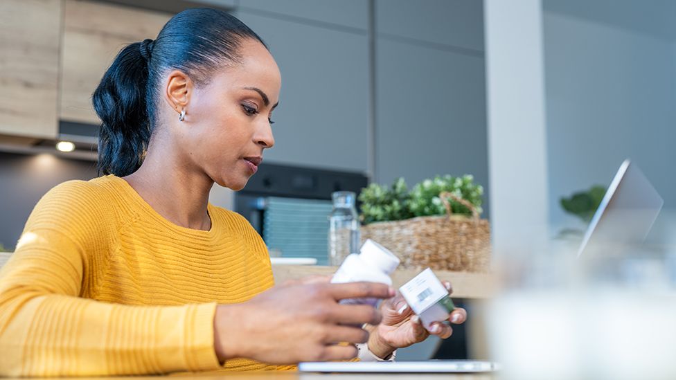 A person wearing a yellow jumper looks at prescription pill bottles while on their computer at home