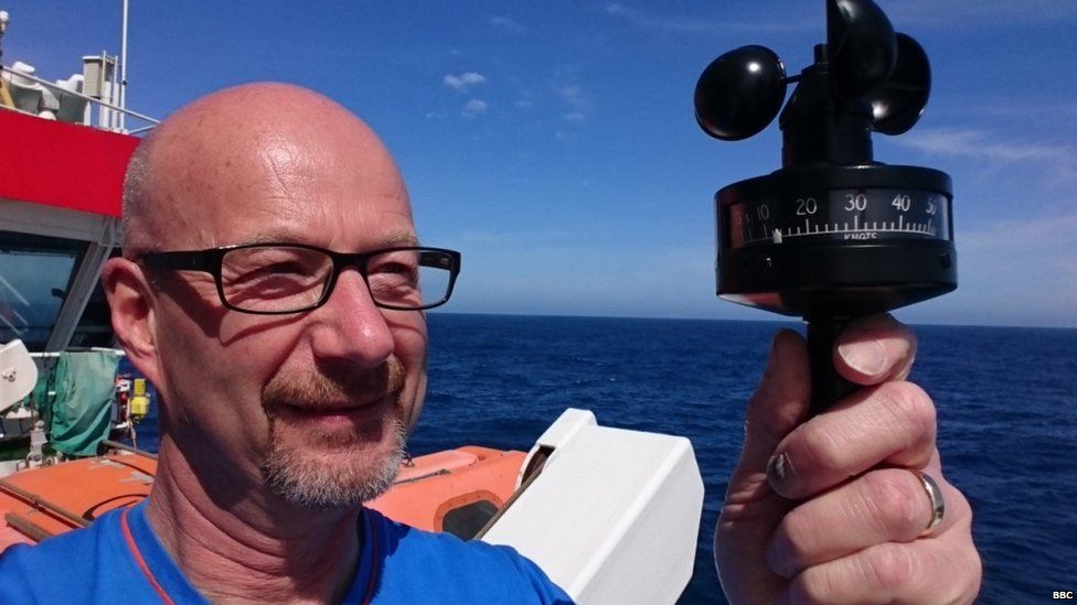 Peter Gibbs onboard holding a black piece of measuring equipment. The sky is blue behind.