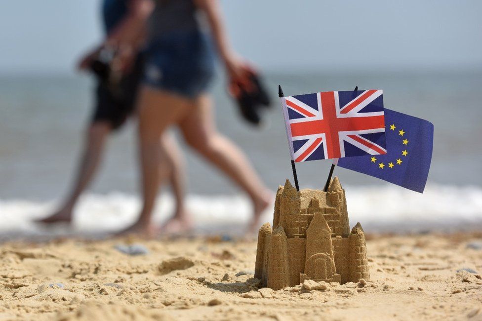 A couple walk on a beach behind a sandcastle with EU and UK flags