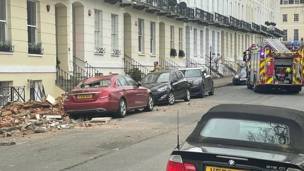 Cars lining a street next to a pavement with rubble on it. A red Mercedes has its rear windscreen smashed.