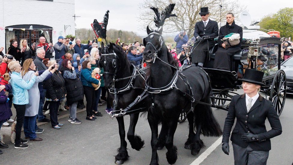 Two black horses pull Paul O'Grady's hearse through Aldington as crowds of mourners, some with dogs, look on