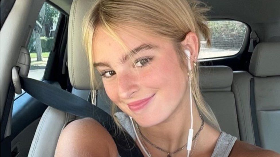 Sophia Rundle, pictured in a selfie taken in a car. Sophia has light blonde hair tied back in a messy bun and hazel eyes - she smiles at the camera with her head tilted over her right shoulder. She has white headphones in her ears and wears a grey vest top and a short necklace. She has her seatbelt pulled over her and behind her a white leather car interior can be seen with a road and greenery visible out of the car windows.