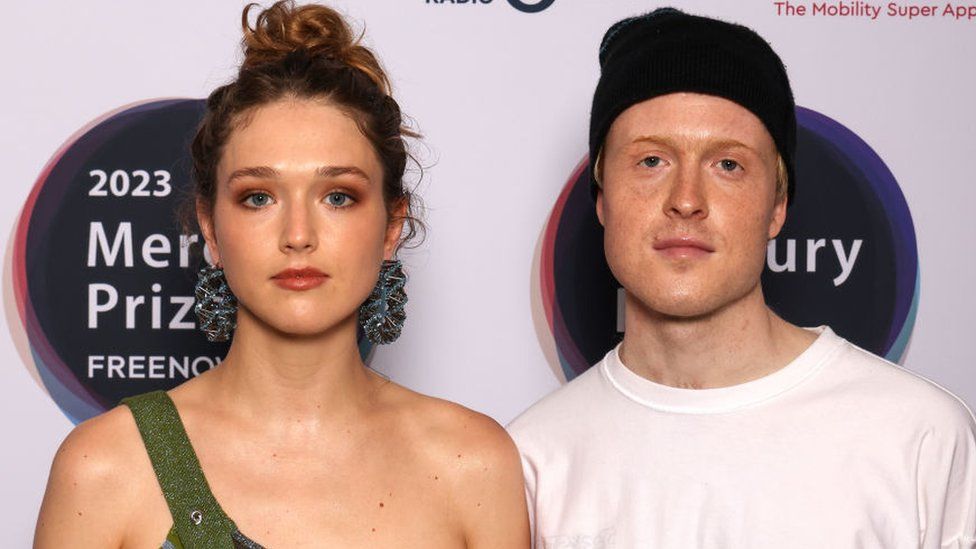 Members of Jockstrap, Georgia Ellery and Taylor Skye attend The Mercury Prize 2023 awards show at Eventim Apollo on September 07, 2023 in London. Georgia stands on the left, she wears a green top over one shoulder and large blue hoop earrings. She has blue eyes and her dark curly hair is tied in a bun on her head. She wears brown-orange eyeshadow and a muted red lipstick. Taylor stands on the right, he has blue eyes, freckled skin and fair hair covered by a dark beanie hat. He wears a white T-shirt and both he and Georgia look at the camera with a neutral expression.