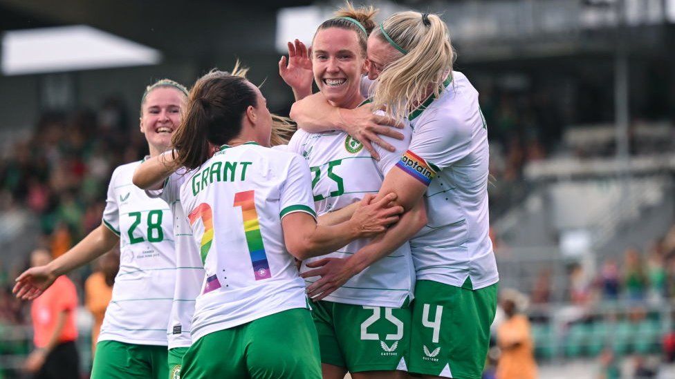 Claire O'Riordan celebrating with her Republic of Ireland team-mates, from near  to close    Erin McLaughlin, Ciara Grant and Louise Quinn aft  scoring a goal. They are each  smiling and wearing their Ireland kit which is achromatic  shirts and greenish  shorts