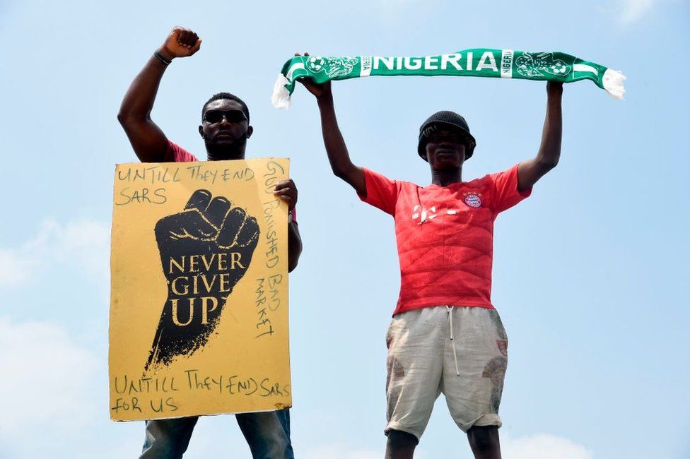 A protester gesture while holding placard as another holds up a scarf with the colours of the Nigerian national flag during a demonstration to protest against police brutality at Magboro, Ogun State in southwest Nigeria, on October 20, 2020. - Authorities declared a 24-hour curfew in Nigeria's economic hub Lagos on October 20, 2020, as violence flared in widespread protests that have rocked cities across the country.