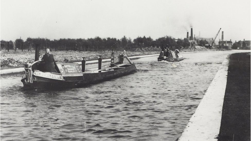 Black and white photograph taken from beside the canal, shows the butty "Sunlight" [built in 1929 by the Anderton Company] in the foreground being towed by the motor "Silver Jubilee" [rebuilt from Anderton Company's horse boat 'Greece']. There is a boatwoman at the tiller of "Sunlight" and a boatman at the tiller of "Silver Jubilee". There are industrial buildings and a crane in the background.