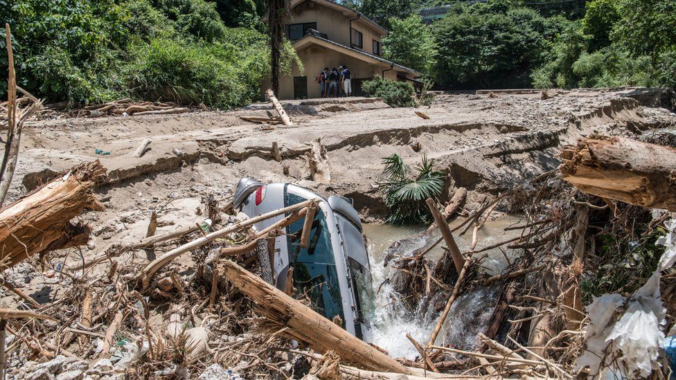 A car lies in mud as people rest in the shade of a house that is partially submerged