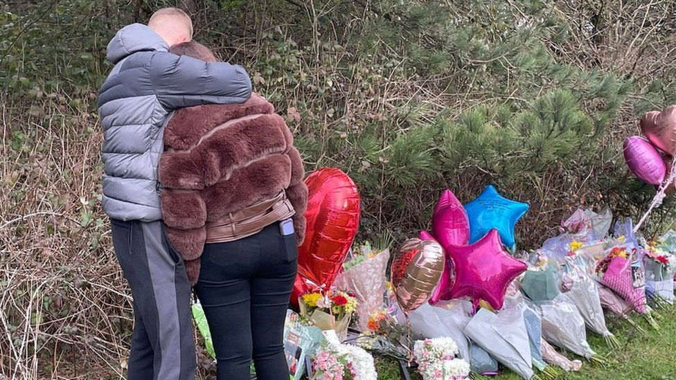 People look at floral tributes at scene of the crash