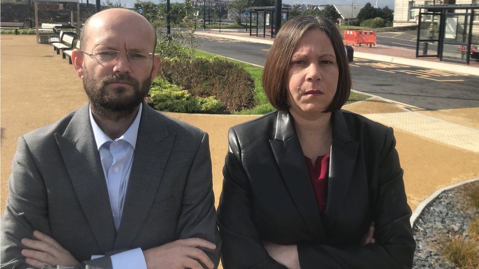 Ian Johnson and Millie Collins standing in front of an empty bus station, both with their arms crossed and looking angry